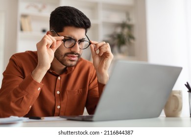 Error. Portrait of confused Arab guy sitting at desk using laptop, taking off glasses looking at pc screen and squinting. Worried man reading bad negative news, having poor eyesight problems