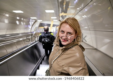 An escalator moves down to an underground metro station, young white woman descends while standing on steps.