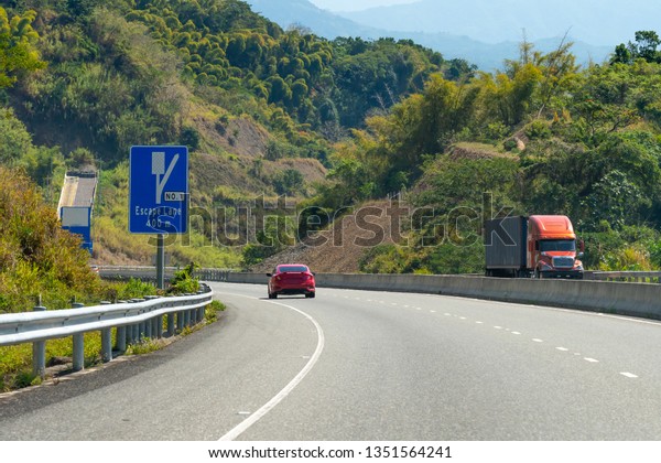 Escape Lane ahead uphill road signs on\
dual carriageway highway through mountains in scenic countryside.\
Red sedan coupe sports car driving on left hand side on two\
lane/double lane\
street/roadway.