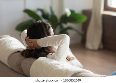 Escaping From Vanity. Happy Tranquil Millennial Woman Spending Free Time Sitting On Couch At Living Room Or In Hotel Suite Alone. Female Putting Hands Behind Head Dreaming Enjoying Watching Tv