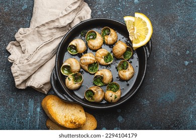 Escargots de Bourgogne Snails with Garlic Butter and Parsley in black cast iron pan with Lemon and Toasted Baguette Slices on rustic stone background top view, traditional French Delicacy .