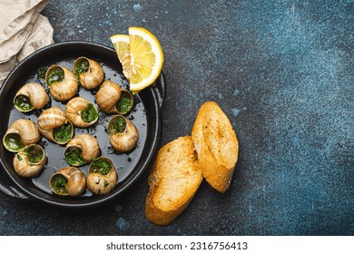 Escargots de Bourgogne Snails with Garlic Butter and Parsley in black cast iron pan with Lemon and Toasted Baguette Slices on rustic stone background top view, traditional French Delicacy, copy space.