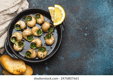 Escargots de Bourgogne Snails with Garlic Butter and Parsley in black cast iron pan with Lemon and Toasted Baguette Slices on rustic stone background top view, traditional French Delicacy, copy space.