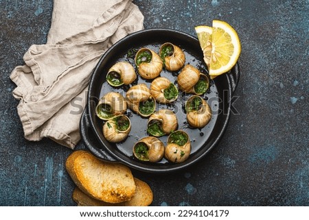 Escargots de Bourgogne Snails with Garlic Butter and Parsley in black cast iron pan with Lemon and Toasted Baguette Slices on rustic stone background top view, traditional French Delicacy .