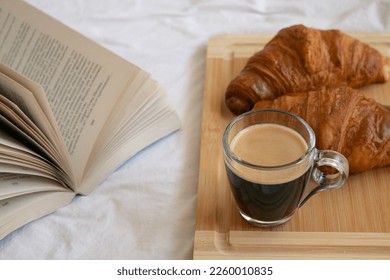 Espresso coffee in a glass mug on a wooden board with croissants and an open book on the bed. Holiday morning - Powered by Shutterstock