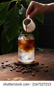 Espresso tonic in the making. Adding coffee into a highball glass filled with ice cubes and tonic soda water, coffee beans, milk jug, wooden background