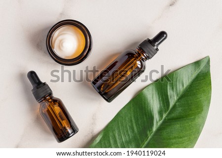 Essential oils with drops pipette and cream on marble background. Grean leaves decoration. Professional bottle for facial and body treatment