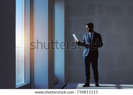 Estate agent stands with tablet computer near big window in blank new apartment on grey wall background. Business concept