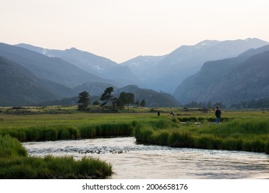 Estes Park, Colorado, USA - July 11, 2021.  Men In Nature Setting With Fly Fishing Rod And A Fast Flowing Stream.  Rocky Mountain Stream With Blue Water, Green Grass