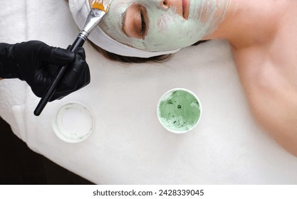 An esthetician gently applies a green, potentially moisturizing, cleansing or nourishing facial mask to a client's face using a brush during a spa session. Horizontal photo - Powered by Shutterstock