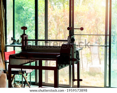 Etching press, art equipment for printmaking. Old red etching press in art studio near glass sliding door on the green garden background outside the building.