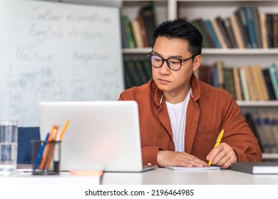 E-Teaching. Korean Middle Aged Man Teacher Using Laptop And Taking Notes Preparing For Virtual Lecture Sitting At Desk In Modern Classroom. Internet Technology And Education. Selective Focus - Shutterstock ID 2196464985