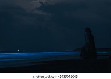 Ethereal blue waves illuminate a night beach, with a solitary rock formation standing under a moonlit sky. - Powered by Shutterstock