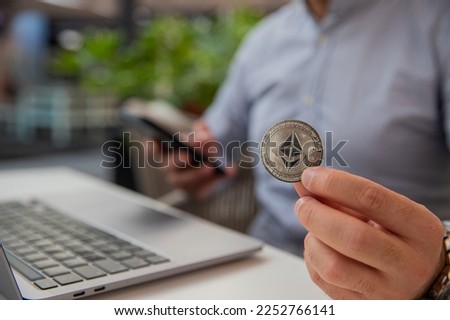 Ethereum coin held in the hand of a trader monitoring the market with his laptop and smartphone