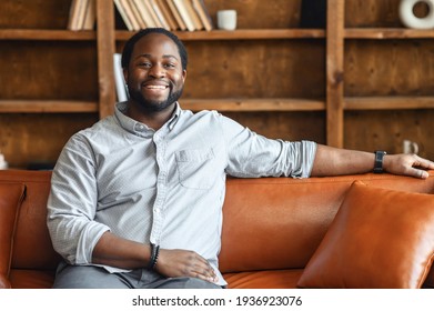 Ethnic black unshaved plump man with dreadlocks sitting on the leather sofa and smiling, in a normal blue shirt and jeans, feeling comfort and relaxation, at an interview, bookcase in the background