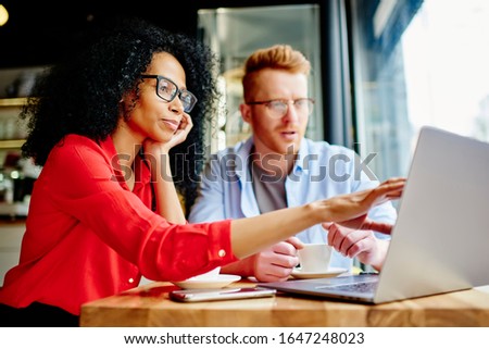ethnic businesswoman in glasses and elegant vibrant red shirt pointing finger on laptop screen and discussing work with pensive colleague while sitting at table and having hot drink in coffee shop