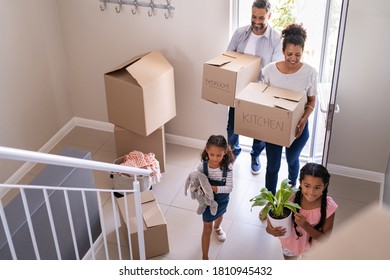 Ethnic family with two children carrying boxes and plant in new home on moving day. High angle view of happy smiling daughters helping mother and father with cardboard boxes in new house.