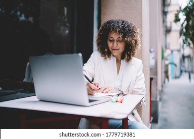 Ethnic Female Freelancer In Casual Outfit And Filling Out Schedule In Daily Planner While Working Remotely On Netbook In Street Cafe