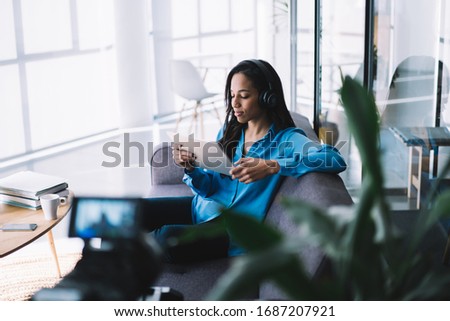 Ethnic female with long dark hair in headphones listening to music and looking at tablet screen while being recorded on camera in modern apartment