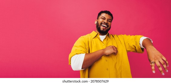 Ethnic man enjoying listening to his favourite music in a studio. Happy young man dancing while streaming music on wireless earphones. Smiling young man standing alone against a pink background.