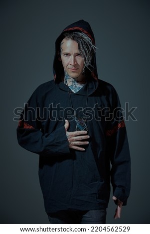 Ethnic rock and punk culture. Portrait of a brutal man with ethnic tattoos and mohawk dreadlocks on his head, posing in black clothes with a hood on a dark grey studio background. Rock musician.