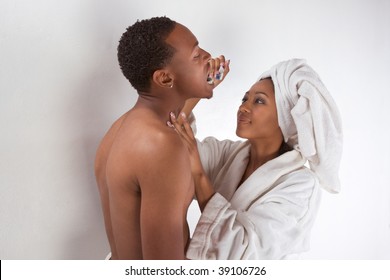 Ethnic Sensual Couple, African American Man And Woman Of Creole Ethnicity Male Wrapped In Bathtowel And Female In White Bathrobe Brushing Teeth Of Her Boyfriend