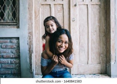 Ethnic small curly girl hugging happy young Indonesian woman from behind while sitting on street with aged door and smiling happily at camera