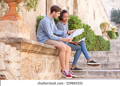 Ethnically Diverse Couple Sitting On A Stone Wall In A Monument Sight Seeing Destination City Sharing A Map On A Summer Holiday, Recreation Outdoors. Travel Lifestyle, Tourist Young Man And Woman.