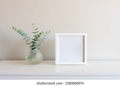 Eucalyptus Leaves In Small Round Glass Vase Next To Blank Square Picture Frame On White Shelf Against Neutral Wall Background