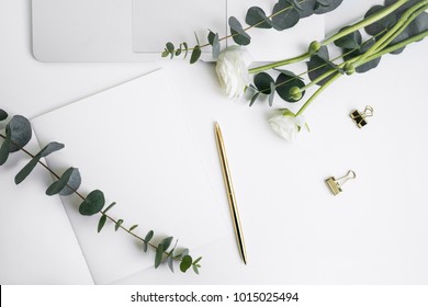 Eucalyptus and ranunculus flower, laptop keyboard, pen, grey copybook with paper mock-up on white table top view. Plants in home office. Feminine floral desktop.