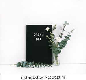 Eucalyptus and ranunculus flower, letter board with motivational quote "Dream big"  on white table. Feminine home office. Minimal floral desktop with white wall.