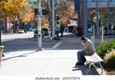 Eugene, OR, 10/29/19: An Older Man On A Bench Looks At His Phone While A Cowboy Sings On The Street Corner And A Woman In Giraffe Pajamas Watches On A Clear Fall Day