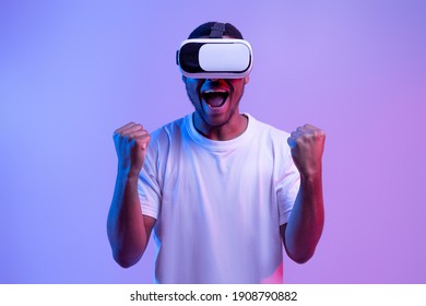 Euphoric Black Guy Playing Video Game In VR Glasses, Emotional African American Man Celebrating Win And Exclaiming With Excitement, Exploring Cyberspace Activities, Neon Light, Purple Background