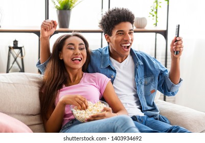 Euphoric Black Teen Couple Watching Tv, Sitting On Couch With Popcorn At Home