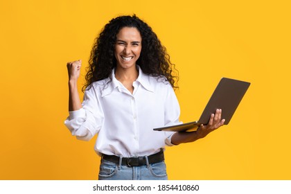 Euphoric Brunette Woman Celebrating Success With Laptop, Raising Clenched Fist, Joyful Millennial Lady Holding Computer, Cheering Victory And Success, Standing On Yellow Studio Background, Copy Space