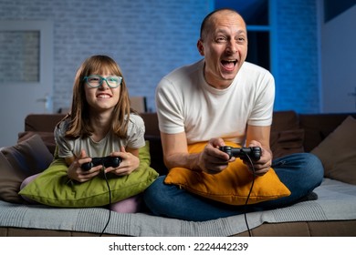 Euphoric Father And Daughter Playing Video Games, Selective Focus. Happy Family Sitting On A Sofa And Playing Video Games With Blue Neon Light In Background. Smiling Family Sitting On The Couch