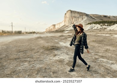 Euphoric woman in hat raises arms in celebration while walking through expansive desert landscape - Powered by Shutterstock