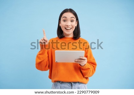 Eureka, got an idea. Excited asian woman with digital tablet, raising finger up, got revelation, has great solution, stands over blue background.