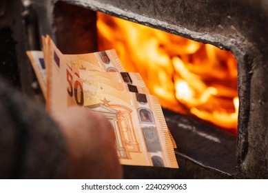 Euro bills in hand of a man near the heating boiler with opened doors and burning flames inside. Spending a lot of money to heat the house, expensive warming
