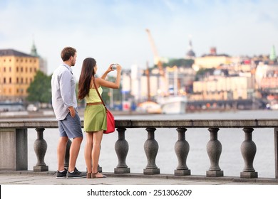 Europe Travel Tourist People Taking Pictures. Tourists Couple In Stockholm Taking Smartphone Photos Having Fun Enjoying Skyline View And River By Stockholm's City Hall, Sweden.