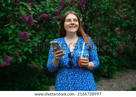 European beautiful woman in bright blue dress holding plastic cold tea cup and looking at smartphone while walking at city park. Businesswoman drinking cold cocktail and smiling. 