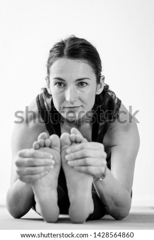 European brunette woman in her mid-thirties folds into a Paschimottanasana or seated forward bend yoga pose, firmly holding her feet and elbows on the floor. Black and white portrait, close-up view.