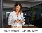 European businesswoman CEO holding smartphone using fintech application standing at workplace in office. Smiling Latin Hispanic mature adult professional business woman using mobile phone cellphone. 