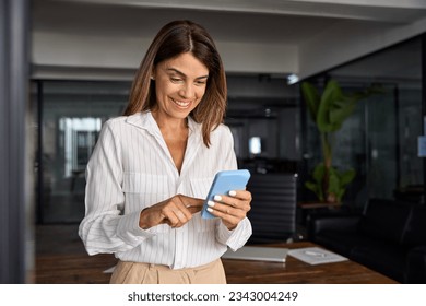 European businesswoman CEO holding smartphone using fintech application standing at workplace in office. Smiling Latin Hispanic mature adult professional business woman using mobile phone cellphone.  - Powered by Shutterstock