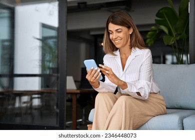 European businesswoman CEO holding smartphone using fintech application sitting on sofa in modern office. Smiling Latin Hispanic mature adult professional business woman using mobile phone cellphone. 