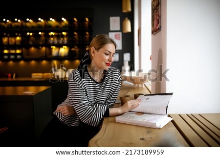 European cute girl in a striped sweater sits and reads a magazine in a cafe. paper cups with coffee. tea. magazine. Expecting friends. Morning mood. natural light. Blonde woman. Kitchen. Home rest