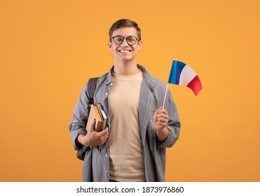 European Education, Modern Study Or Exchange Student. Young Happy Guy In Glasses With Backpack, Notepads And Small Flag Of France, Isolated On Orange Background, Copy Space, Studio Shot