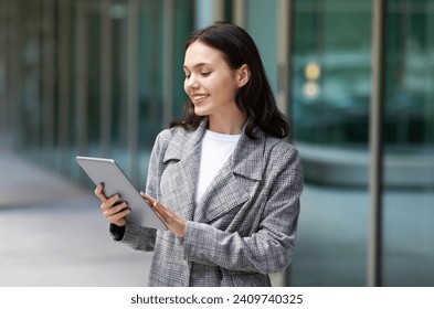 European entrepreneur woman using her digital tablet on city street, smiling while browsing internet and working online in urban area outside. Technology and career success - Powered by Shutterstock