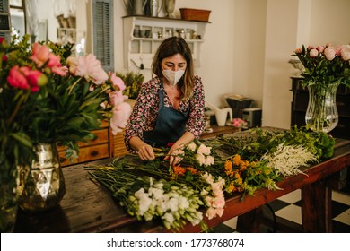 A European female florist wearing a medical face mask and a green apron making flower arrangements in a floral design studio - Covid-19 concept - Powered by Shutterstock