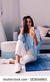 European Fit Brunette Fashion Blogger Woman Sits On Floor In Living Room Near Sofa, Wearing White Jeans And Blue Sweather, And Take Photo Selfie In Mirror   